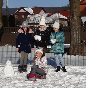 Cztery dziewczynki z klasy III b na szkolnym placu w scenerii zimowej, ubrane w zimowe ubrania, trzy uczennice stoją, jedna przed nimi siedzi na śniegu