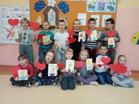 dzieci z klasy 0 b, ustawione w dwóch rzędach, trzymają w rękach serca z papieru i laurki dla babć i dziadków