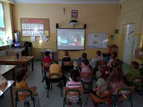 Dzieci z klasy 0 siedzą na krzesełkach, oglądają film na tablicy multimedialnej