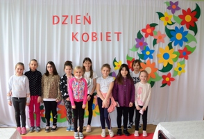 na ścianie napis Dzień Kobiet i kwiaty wokół, przed dekoracją stoją dziewczynki